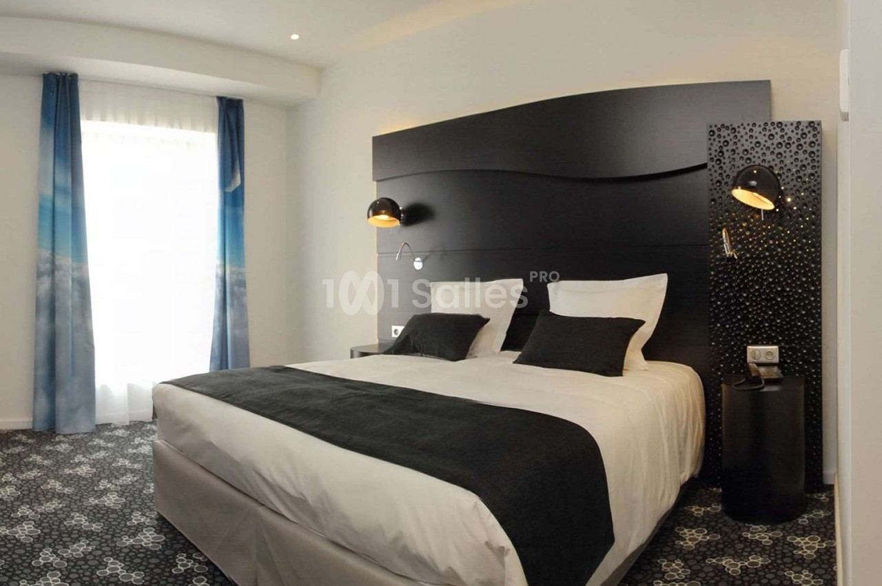 Chambre d'hôtel moderne avec lit double, tête de lit noire, lampes murales et rideaux bleus près d'une fenêtre.