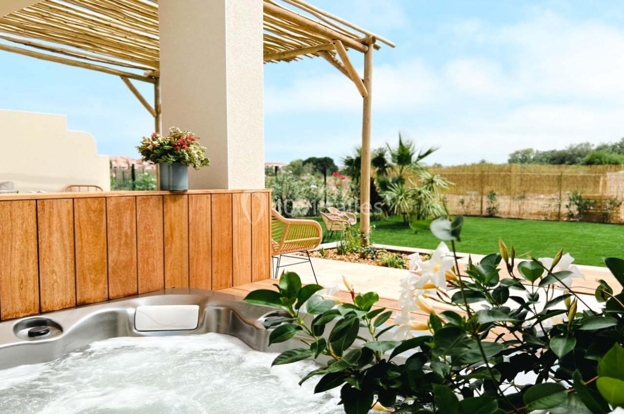 Bain à remous en premier plan sur une terrasse ensoleillée avec pergola, plantes et jardin verdoyant en arrière-plan.