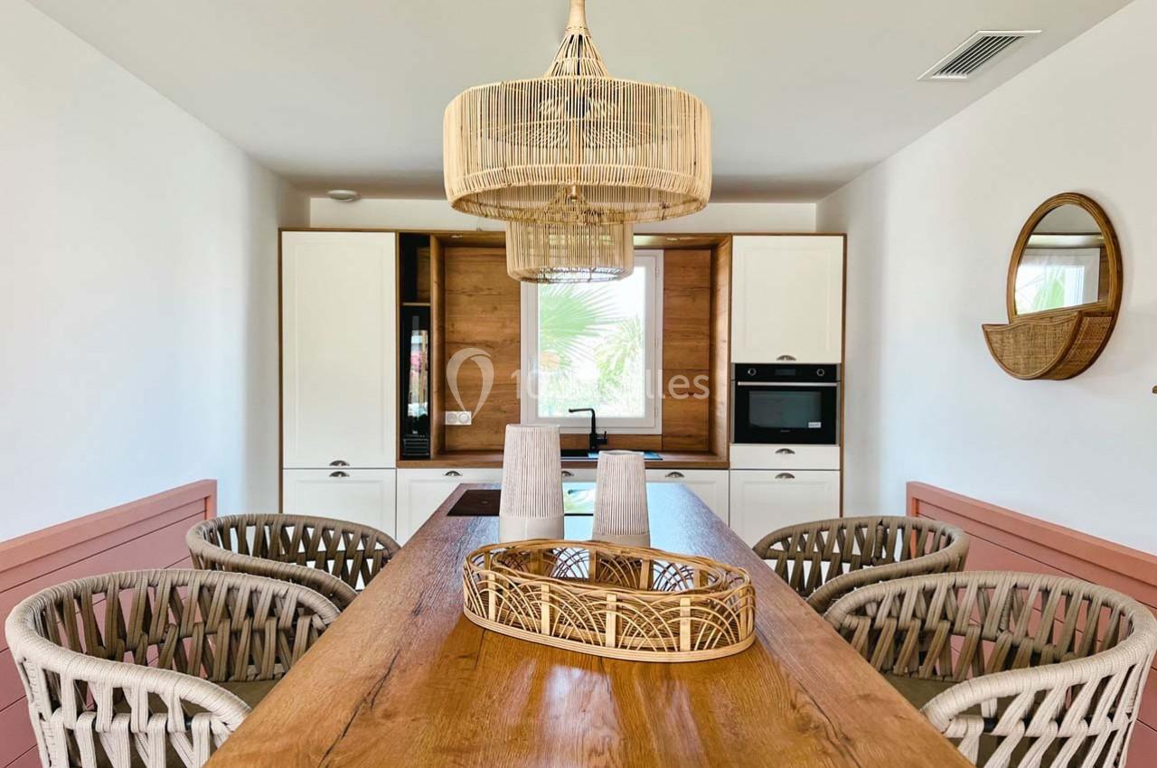 Salle à manger lumineuse avec table en bois, chaises tressées, suspension en rotin et cuisine équipée en arrière-plan.