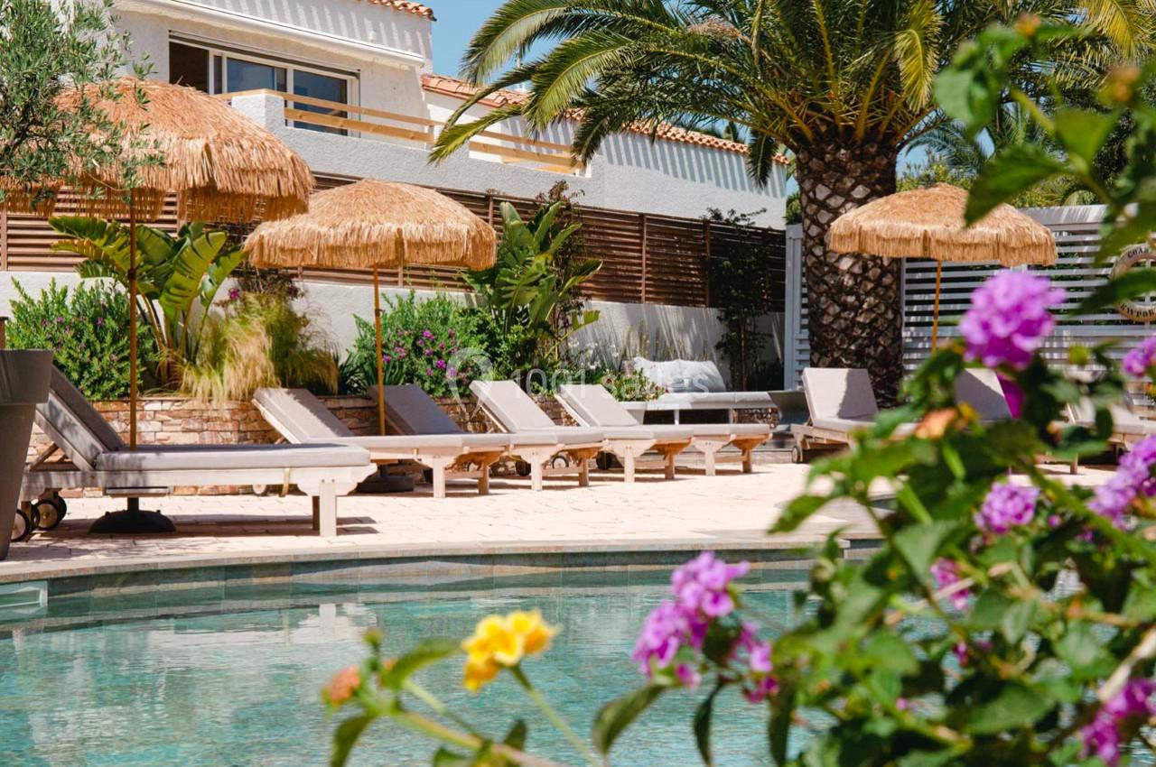 Terrasse avec piscine, transats en bois, parasols en paille, végétation tropicale et maison en arrière-plan.