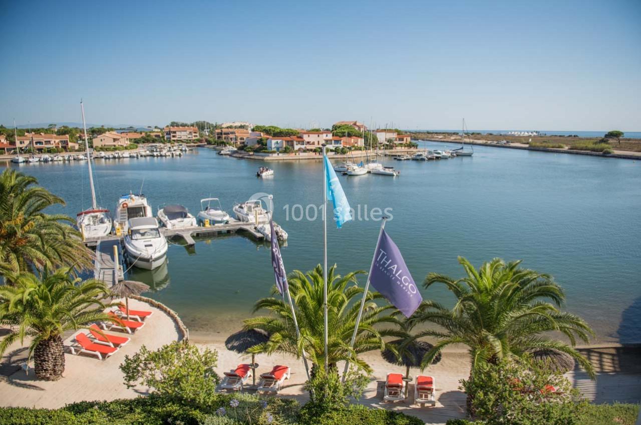 Vue d'un port de plaisance avec des bateaux amarrés, des palmiers et des bâtiments en arrière-plan sous un ciel dégagé.
