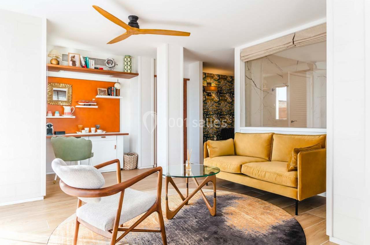 Salon lumineux avec canapé jaune, fauteuils design, table en verre, mur orange et bureau intégré en arrière-plan.