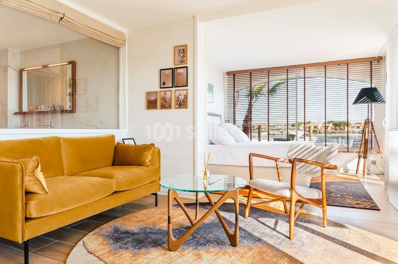 Salon lumineux avec canapé jaune, table basse en verre, fauteuils en bois et vue sur une chambre et l'extérieur.