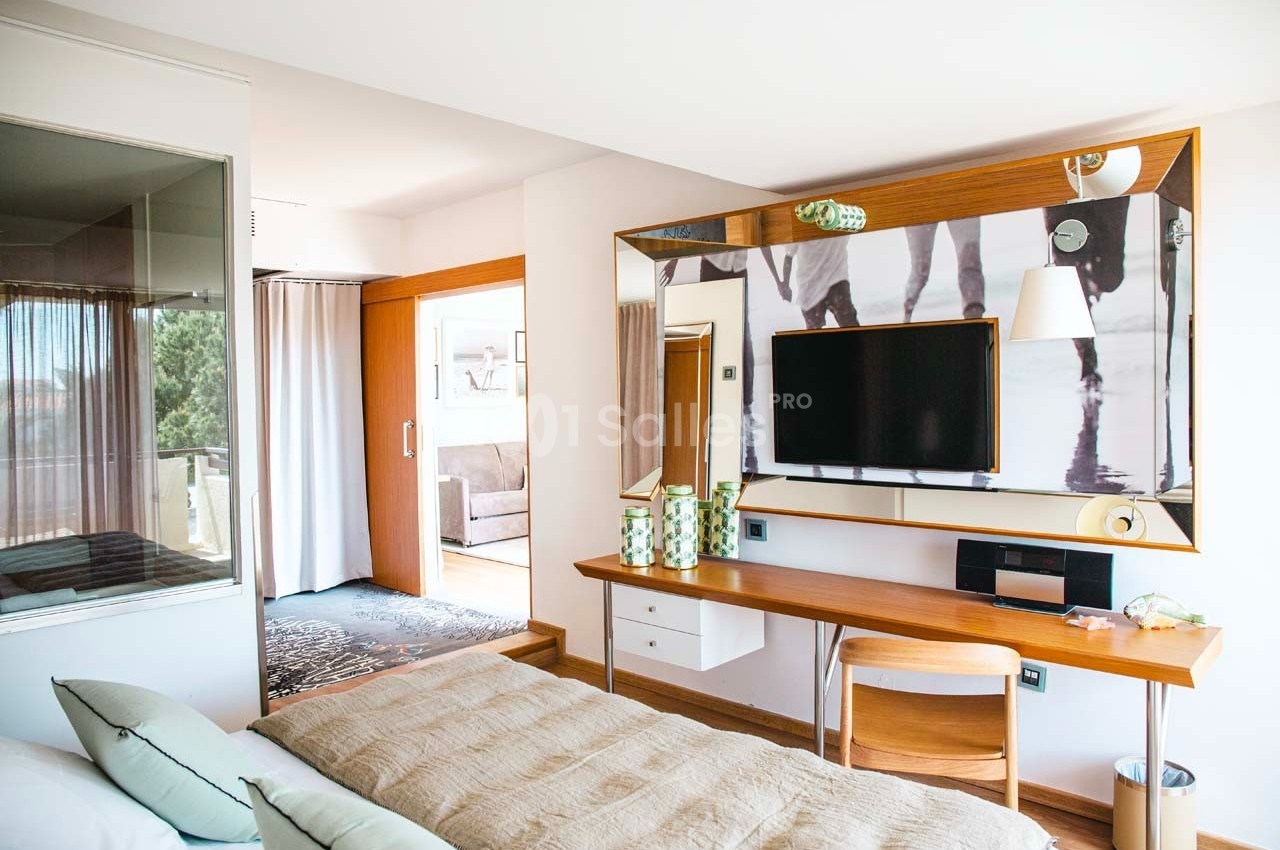 Chambre lumineuse avec lit, bureau en bois, télévision murale et accès à un balcon avec vue extérieure.