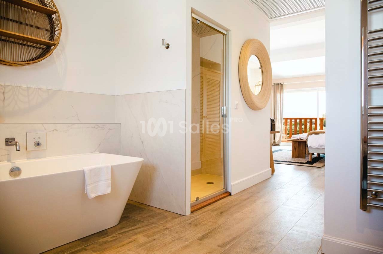 Salle de bain lumineuse avec baignoire blanche, douche vitrée, miroir rond et vue sur une chambre avec lit.