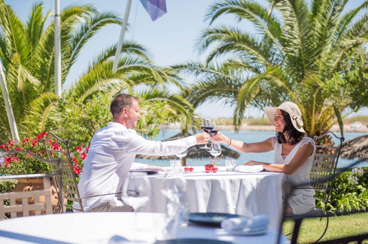 Un couple trinque avec des verres de vin à une table en extérieur, entouré de palmiers et d'un paysage ensoleillé.