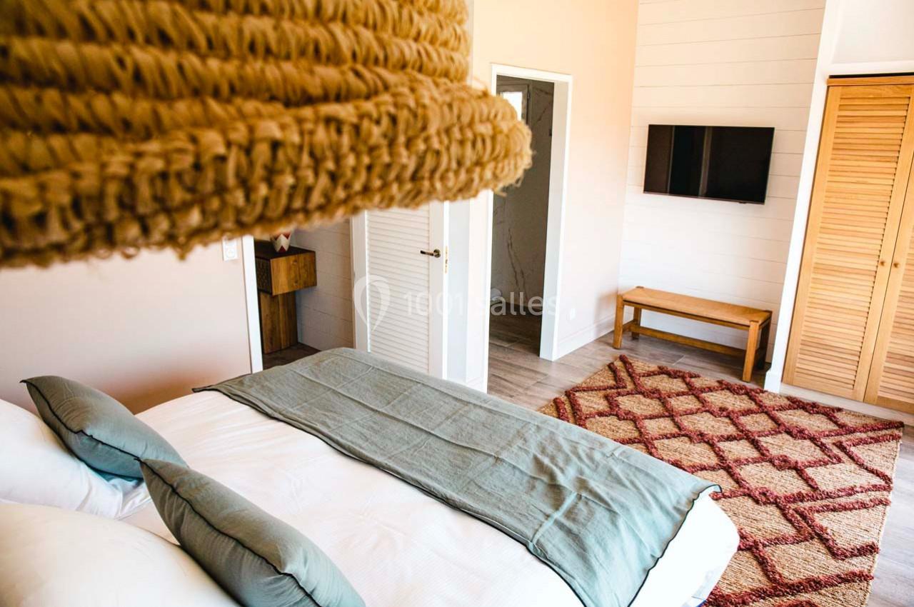 Chambre lumineuse avec lit double, couverture verte, tapis à motifs rouges, télévision murale et portes en bois clair.