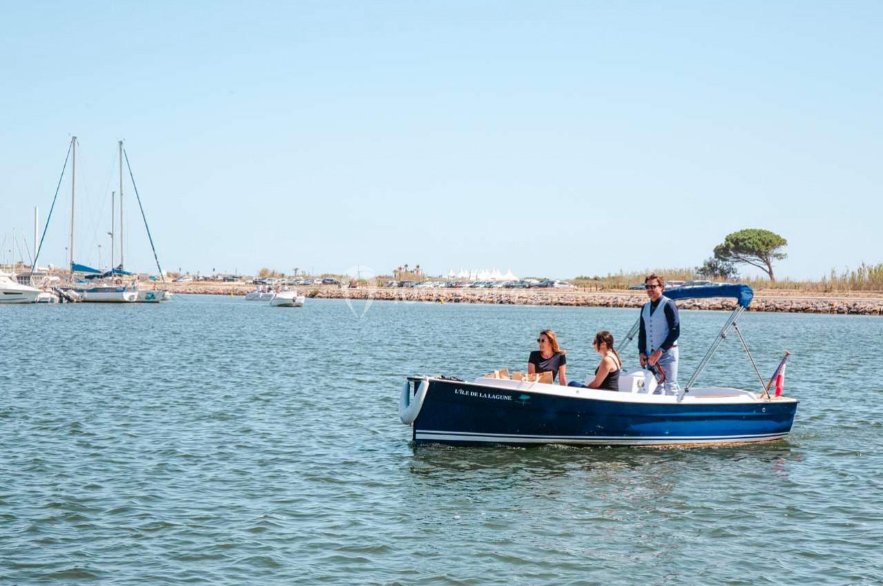Un petit bateau à moteur navigue sur une eau calme avec trois personnes à bord, un port visible en arrière-plan.