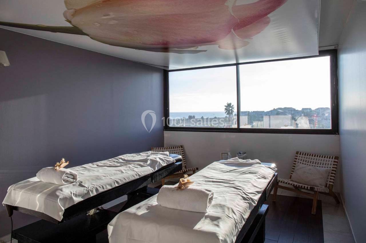 Salle de massage lumineuse avec deux tables de soins, vue sur l'extérieur et plafond décoré d'une fleur.