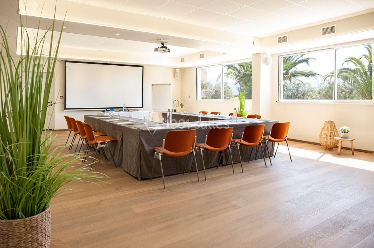 Salle de réunion lumineuse avec grandes fenêtres, table rectangulaire couverte d'une nappe grise et chaises orange.