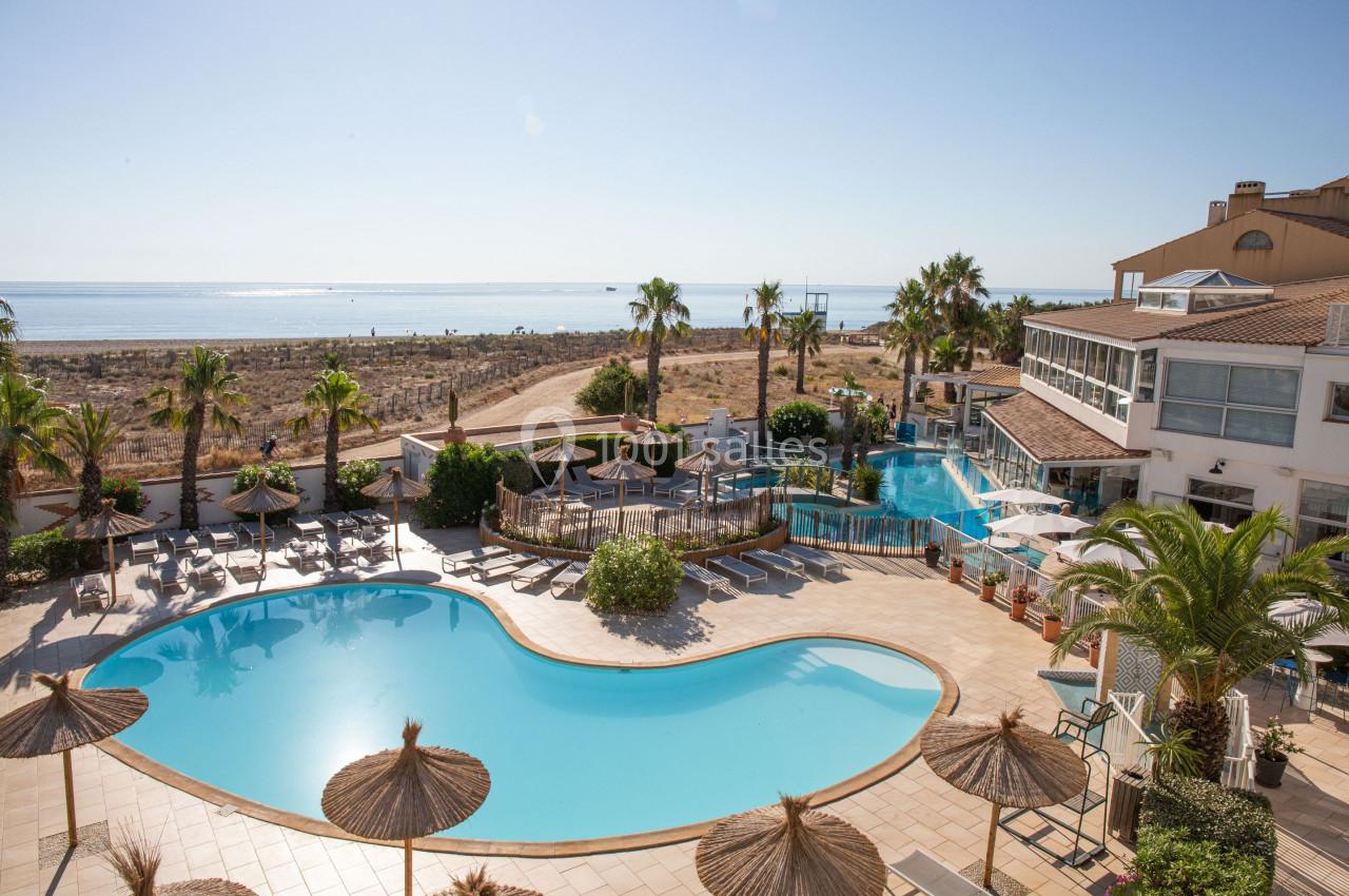 Vue d'une piscine extérieure avec transats, parasols en paille, palmiers et aperçu de la mer en arrière-plan.