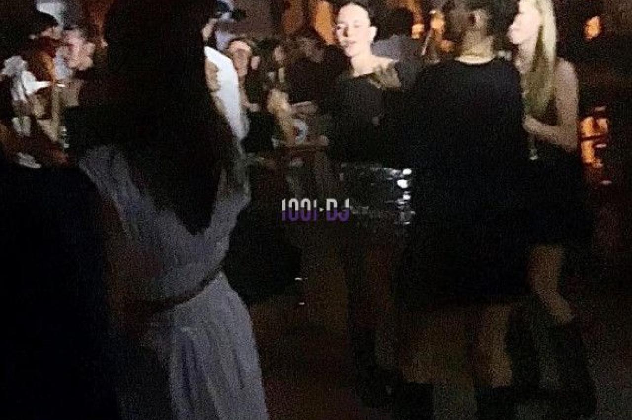 Personnes dans une ambiance tamisée, discutant et dansant près d'un bar décoré de fleurs suspendues.