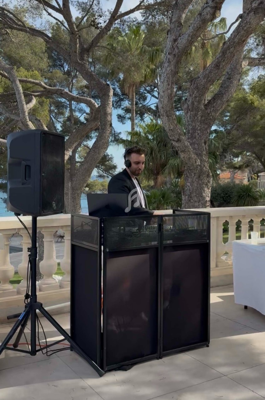 Un DJ en costume mixe de la musique en extérieur, devant une balustrade avec vue sur des arbres et la mer.