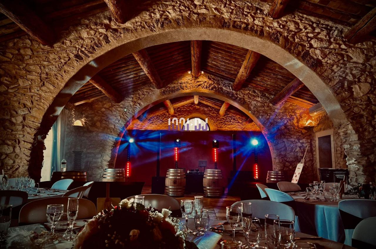 Salle en pierre voûtée décorée pour un événement, avec tables dressées et éclairages colorés sur une scène.