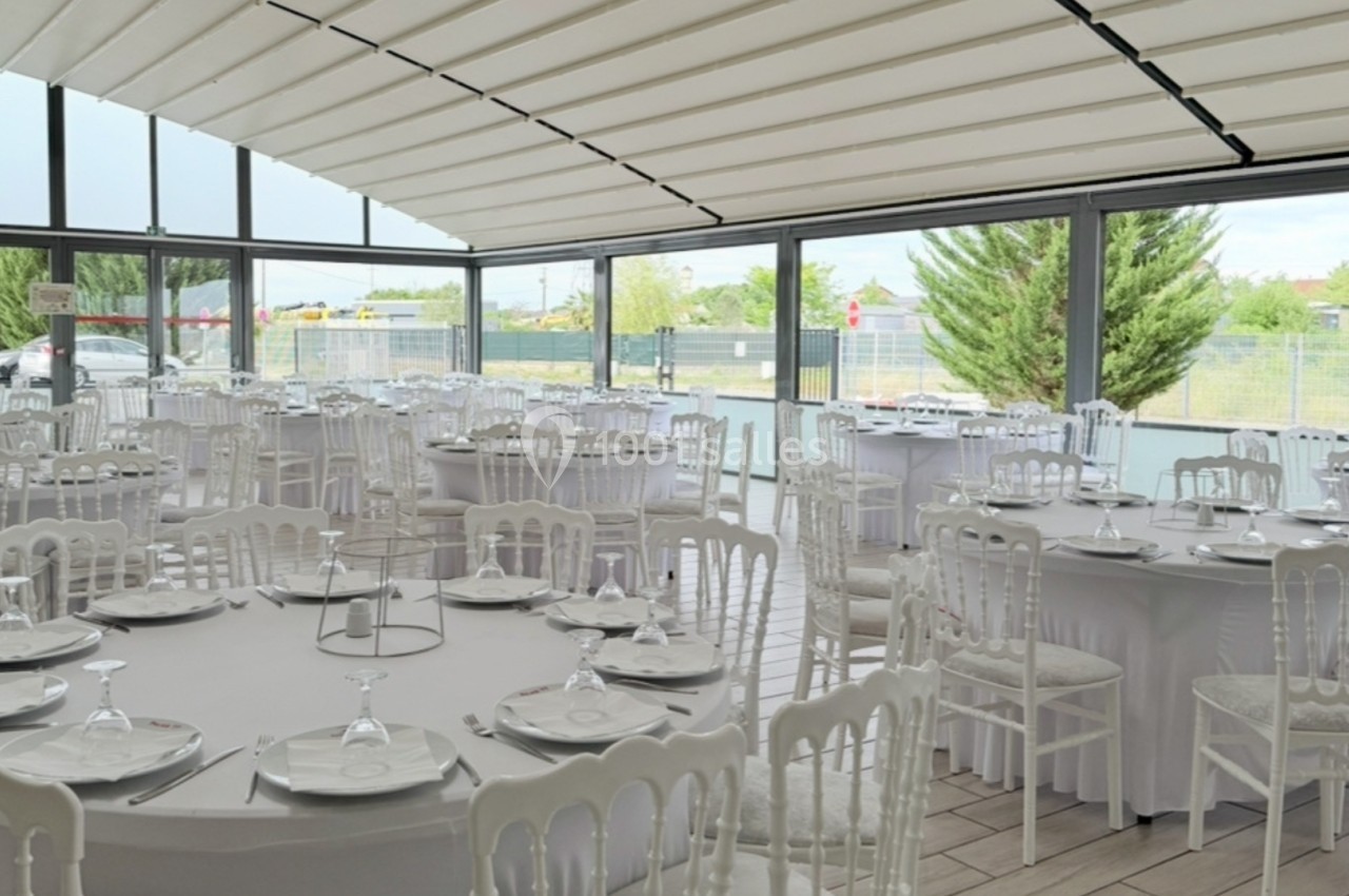 Salle de réception lumineuse avec tables rondes dressées de nappes blanches, entourées de chaises blanches.