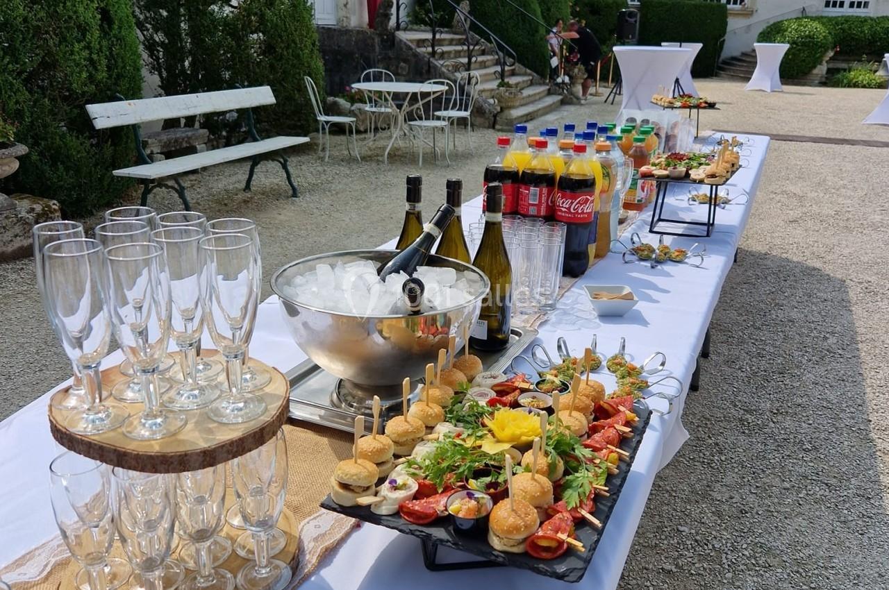 Buffet en extérieur avec verrines, canapés, bouteilles de vin et boissons gazeuses sur une table dressée.