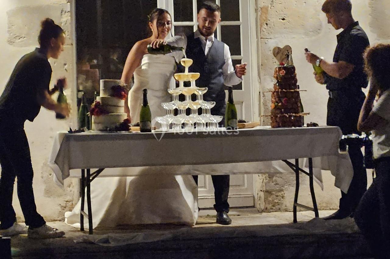 Un couple de mariés devant une pièce montée et des bouteilles de champagne, entouré de personnes en extérieur.