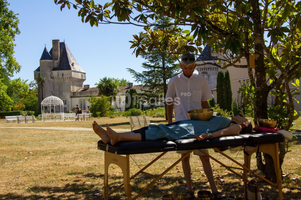 Un homme pratique une séance de relaxation sonore sur une femme allongée, dans un parc près d'un château.