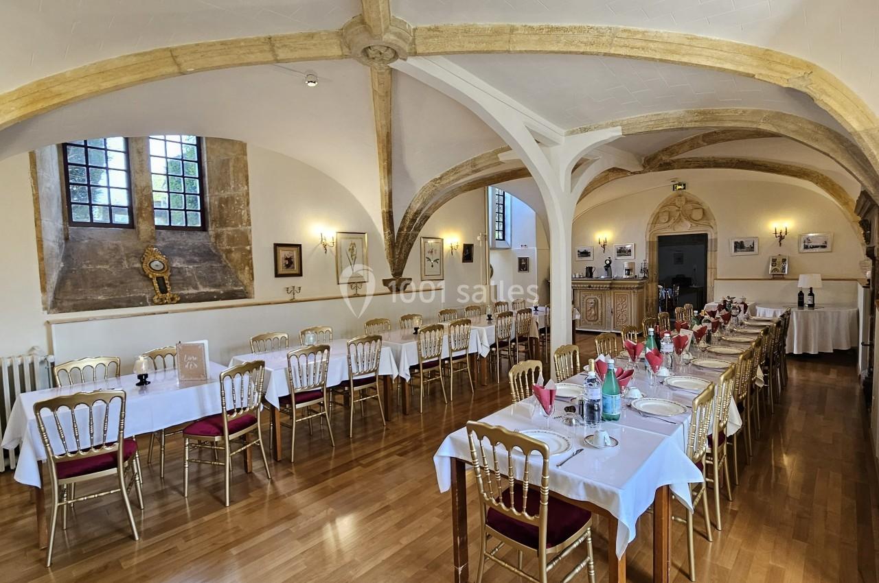 Salle de réception lumineuse avec tables dressées, chaises dorées et poutres apparentes dans un cadre historique.