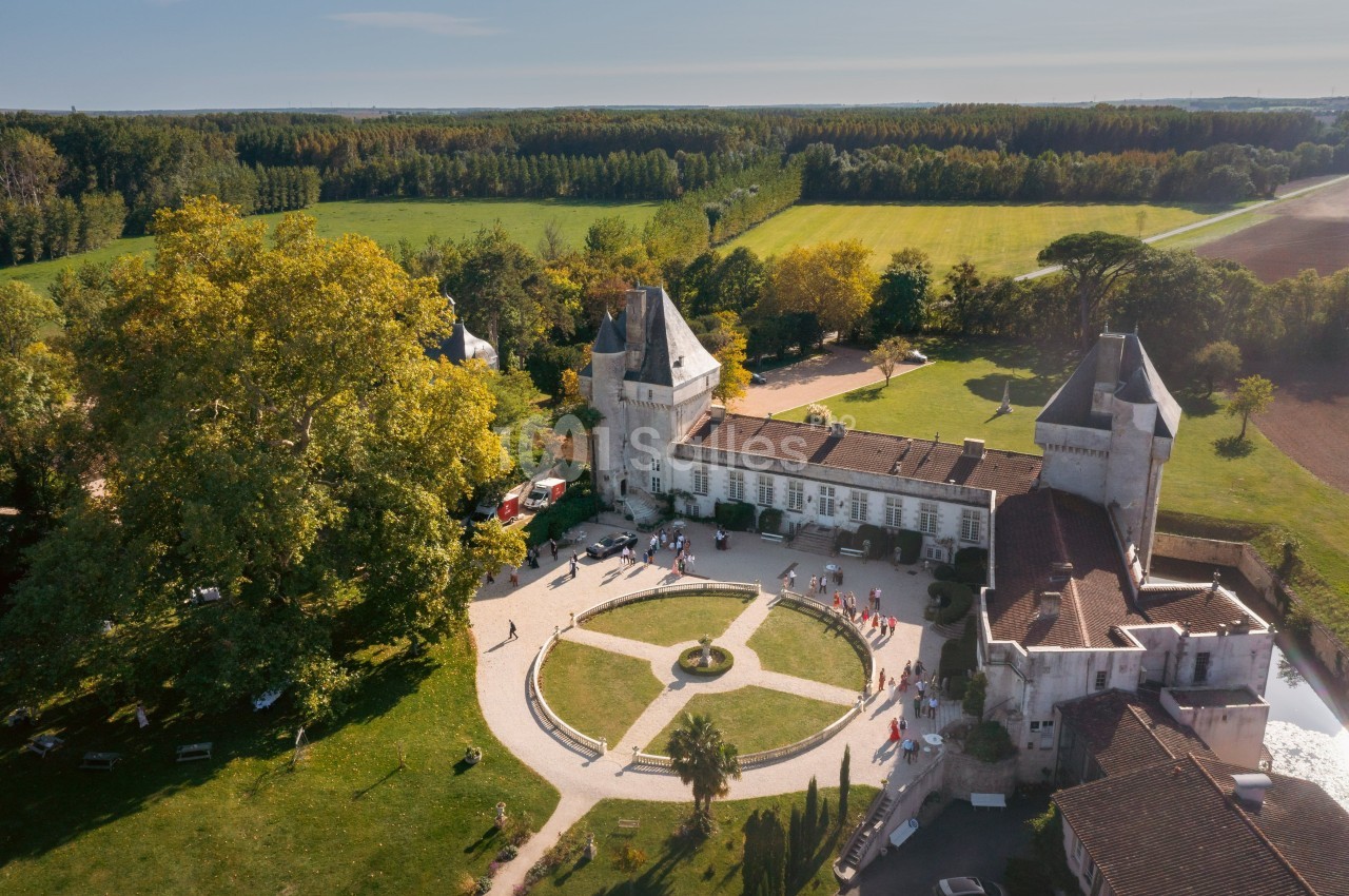 Vue aérienne d'un château entouré de jardins, avec des visiteurs et des champs verdoyants à l'arrière-plan.