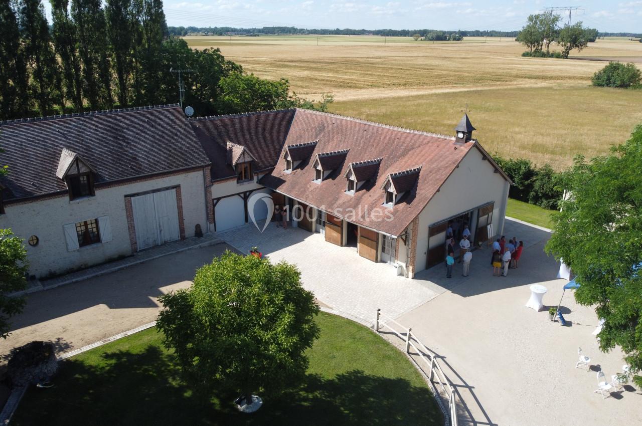 Vue aérienne d'une maison de campagne avec toit en tuiles, entourée de champs et d'arbres, avec des personnes rassemblées.