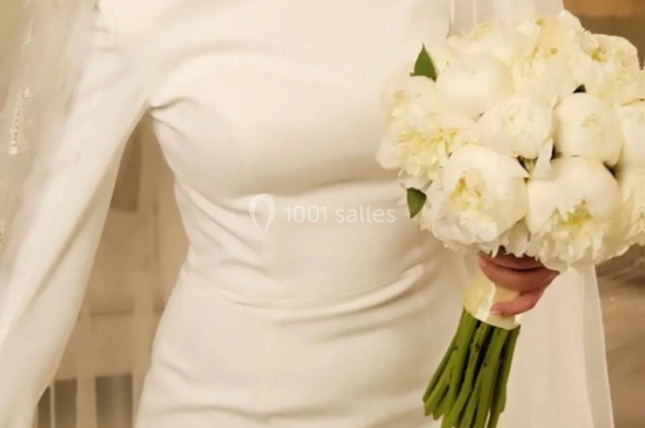 Une mariée en robe blanche tient un bouquet de pivoines blanches dans un décor intérieur lumineux.