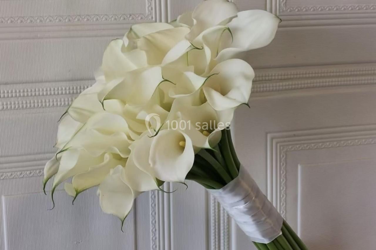 Bouquet de callas blancs élégamment disposés, attachés avec un ruban blanc, posé devant un mur texturé.