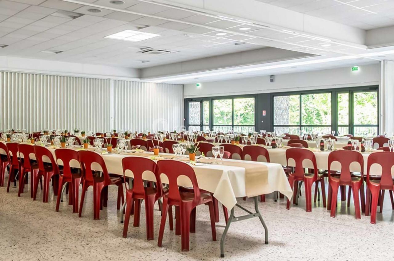 Salle de réception lumineuse avec des tables dressées, nappes blanches et chaises rouges, prête pour un événement.