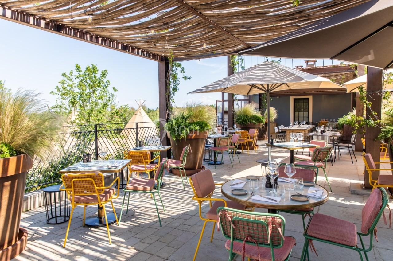 Terrasse ensoleillée avec tables dressées, chaises colorées et pergola en bois, entourée de végétation.