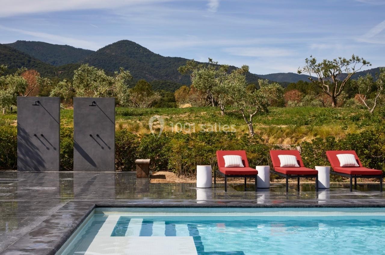 Piscine extérieure avec chaises longues rouges, douches modernes et vue sur des collines verdoyantes.