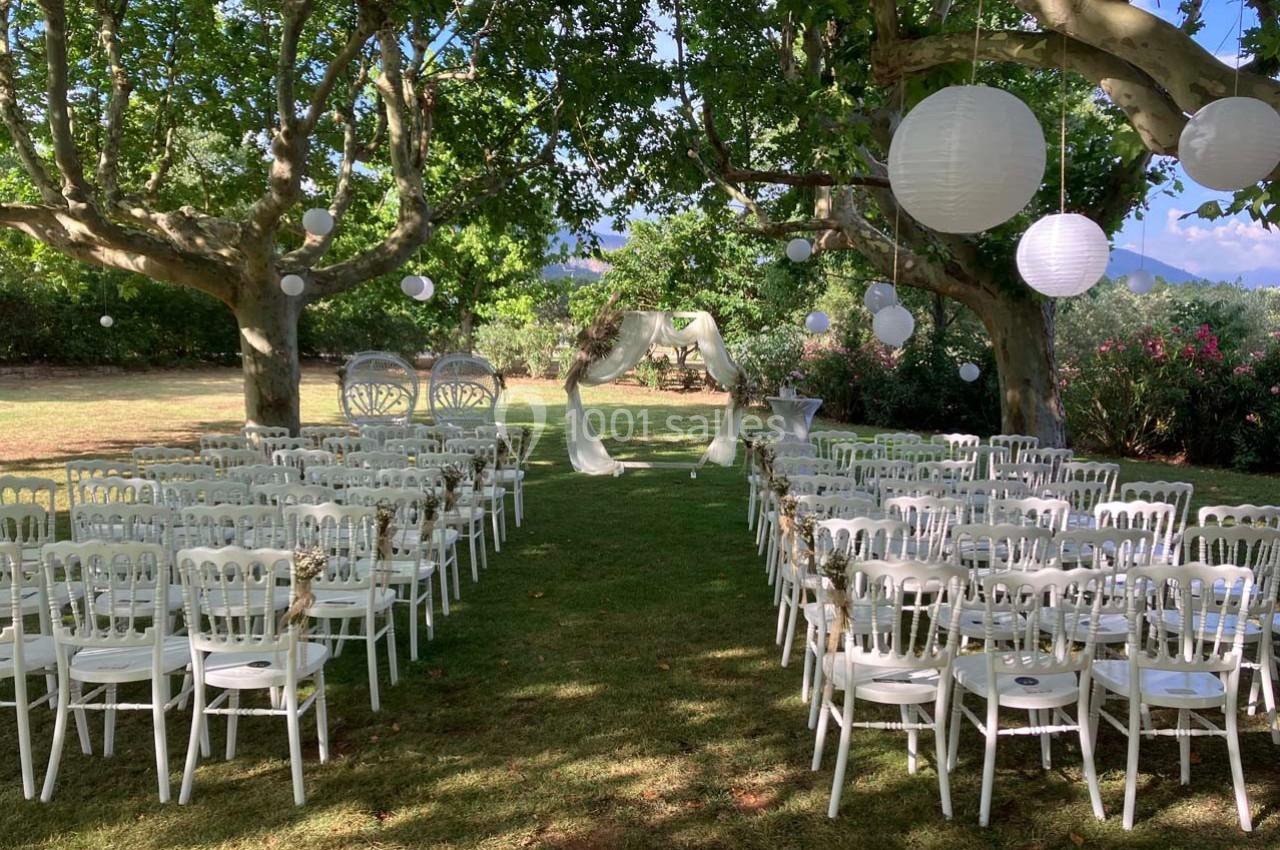 Chaises blanches alignées en extérieur sous des arbres, avec une arche décorée et des lanternes suspendues.