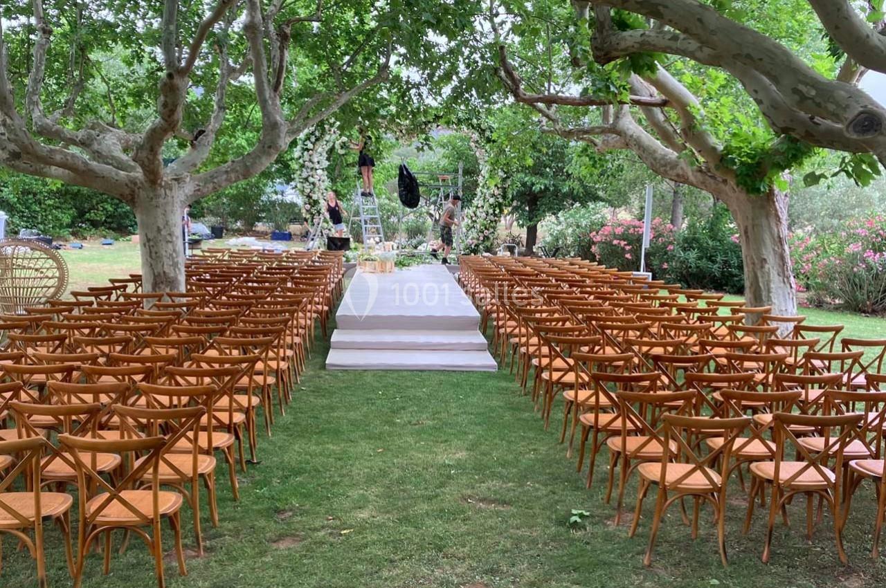 Allée centrale bordée de chaises en bois menant à une estrade décorée pour une cérémonie en plein air sous des arbres.