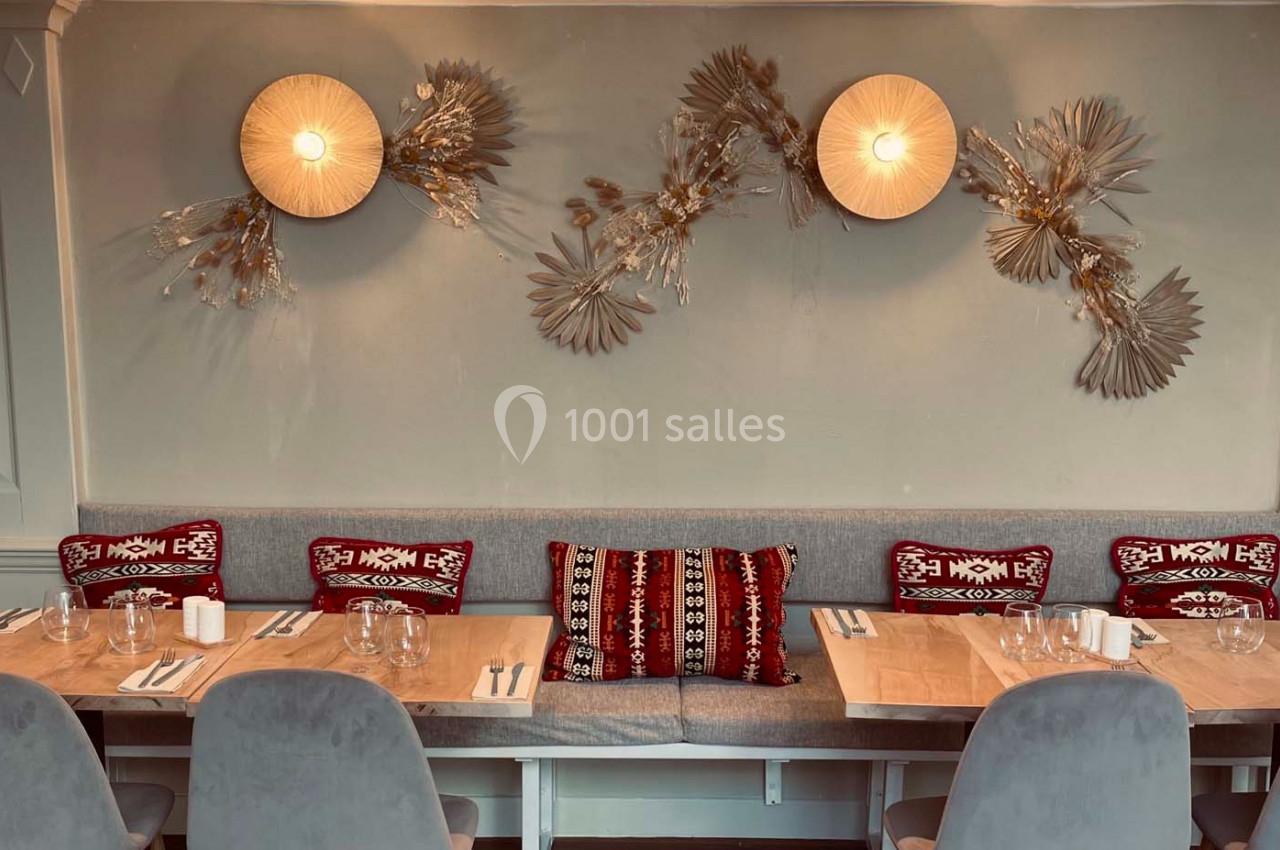 Banquette grise avec coussins rouges à motifs, tables en bois dressées, décor mural avec luminaires et ornements végétaux.