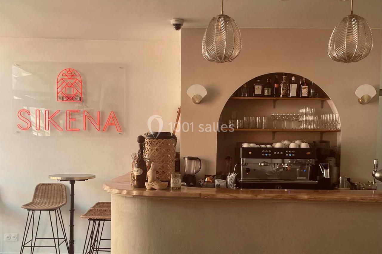 Bar avec comptoir en bois, machine à café, étagères avec bouteilles et verres, et enseigne lumineuse ’Sikena’.