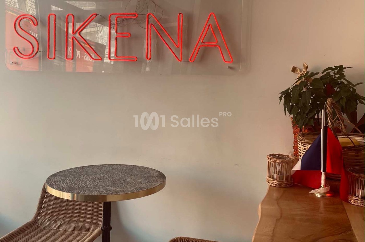 Enseigne néon rouge ’SIKENA’ sur un mur blanc, avec une table, une chaise et un drapeau sur un comptoir en bois.