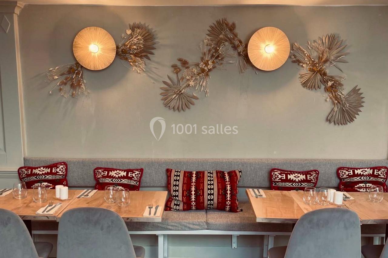 Banquette grise avec coussins rouges à motifs, tables en bois dressées et décoration murale florale sous des appliques…