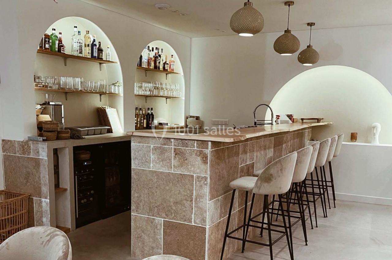 Bar moderne avec comptoir en pierre, tabourets hauts, étagères éclairées contenant des bouteilles et des verres.