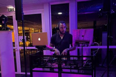 Un DJ souriant derrière une table équipée de matériel audio, avec un ordinateur portable et une table de mixage.