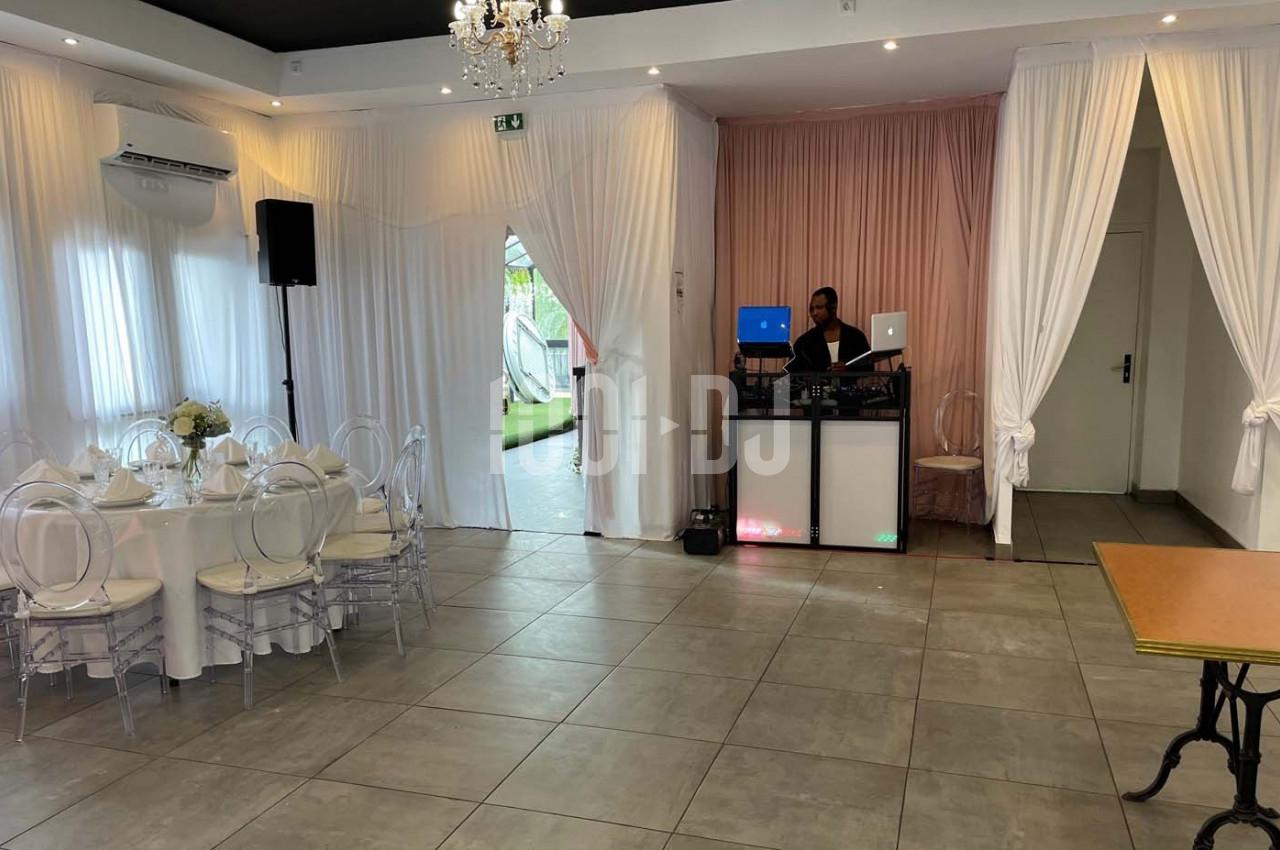 Salle de réception lumineuse avec table dressée, rideaux blancs et DJ installé près d'un mur rose.