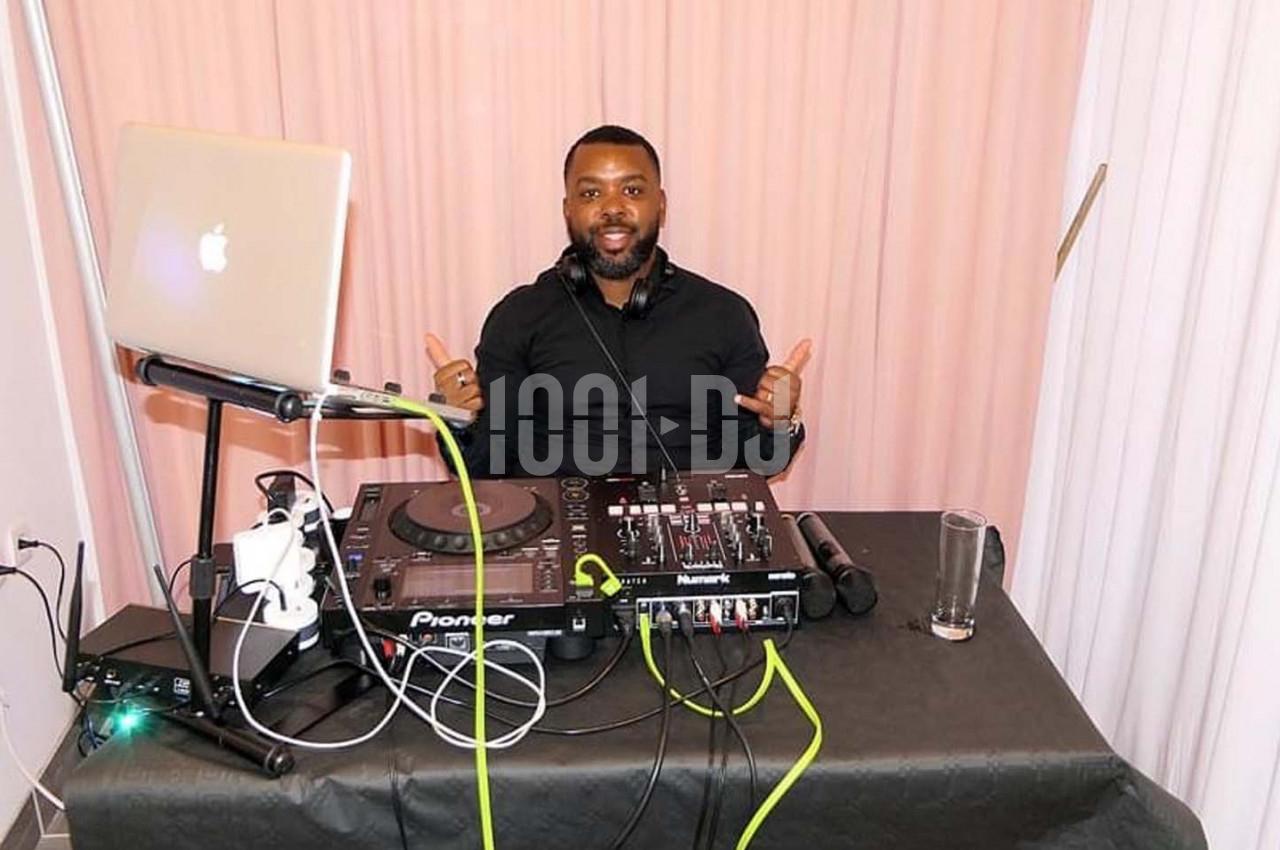 Un DJ souriant derrière une table de mixage avec un ordinateur portable et des équipements audio.