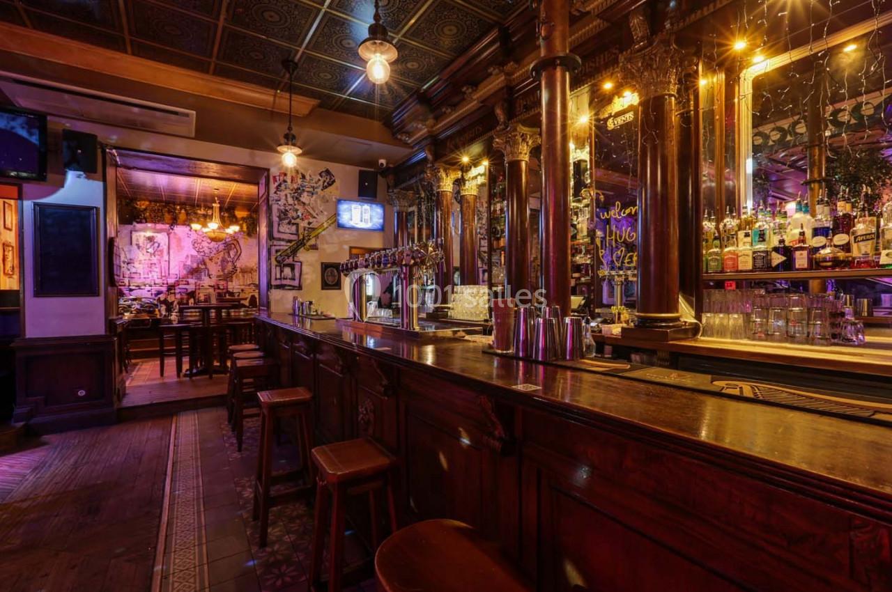 Intérieur d'un bar chaleureux avec comptoir en bois, tabourets, étagères de bouteilles et éclairage tamisé.