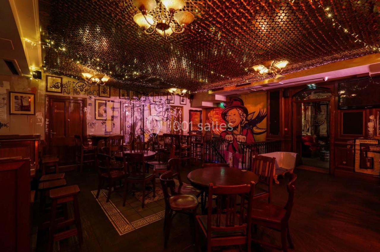 Salle de bar chaleureuse avec tables en bois, éclairage tamisé, fresque murale colorée et filet décoratif au plafond.