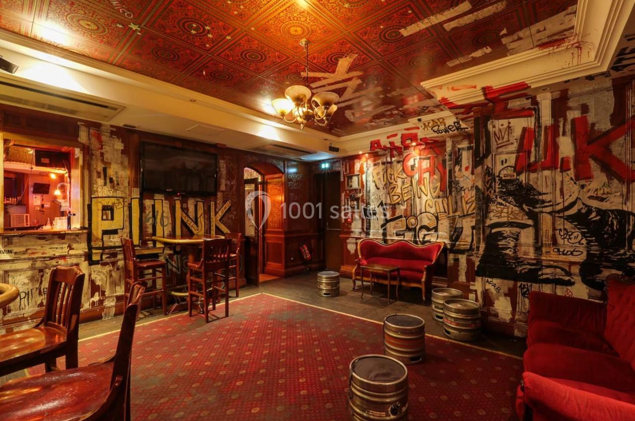Salle de bar à la décoration punk avec murs graffités, tonneaux métalliques et mobilier en bois et velours rouge.