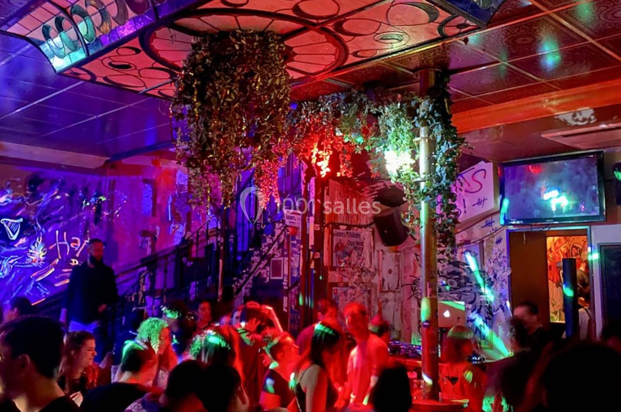 Salle animée avec des personnes dansant sous des lumières colorées, décorée de graffitis et de plantes suspendues.
