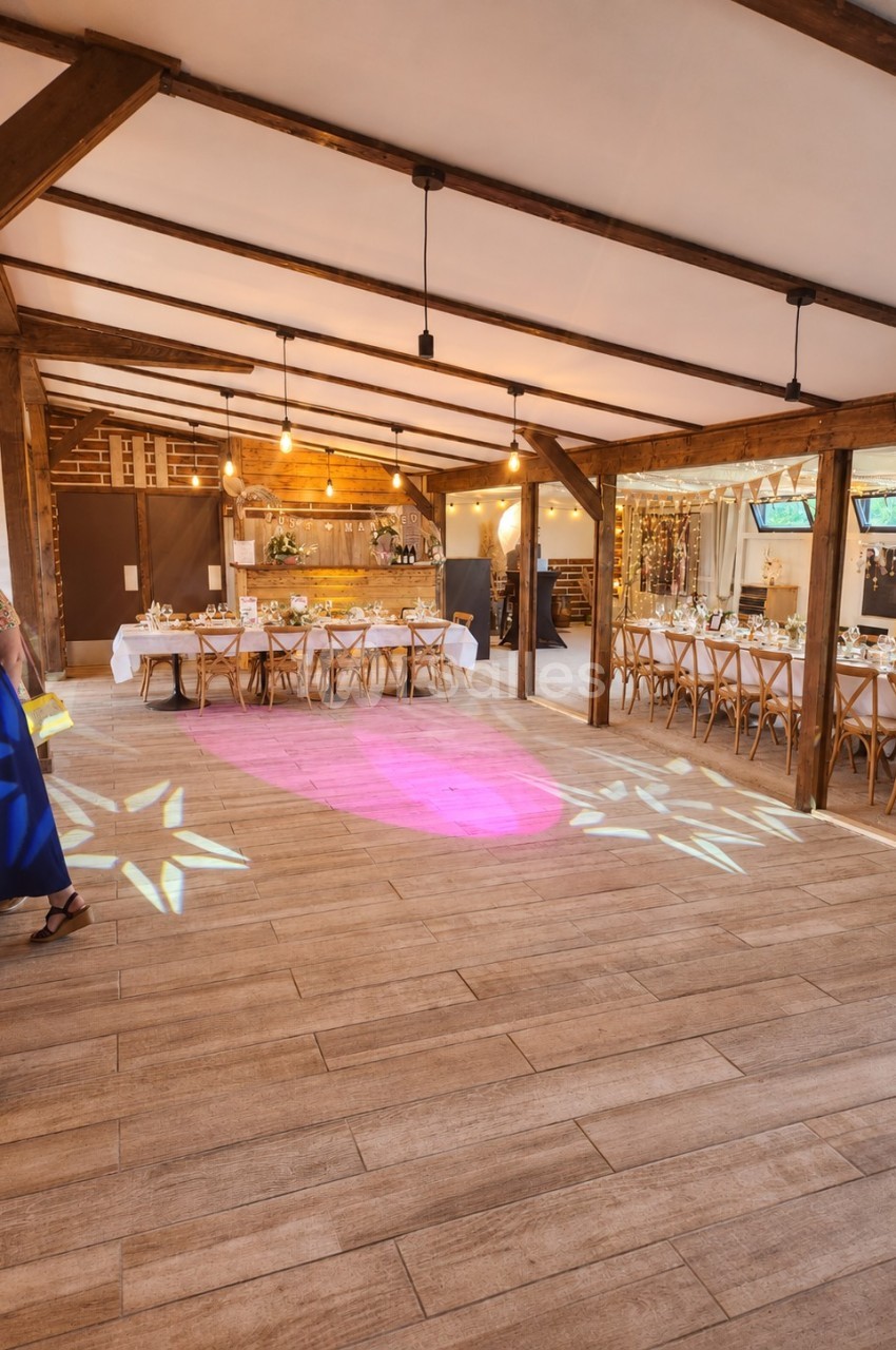 Salle de réception avec poutres apparentes, tables dressées, éclairage suspendu et piste de danse rose illuminée.
