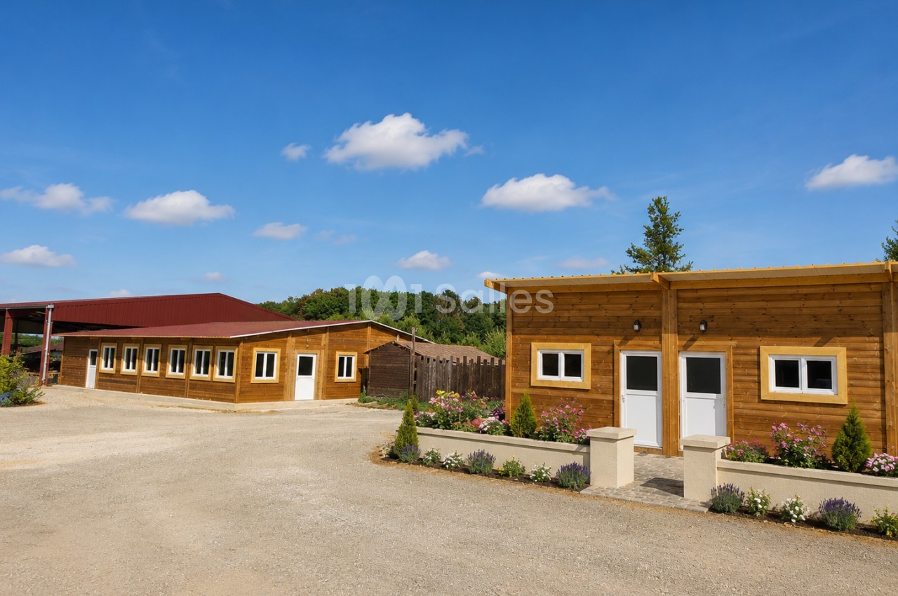 Ensemble de bâtiments en bois avec toiture rouge, allée gravillonnée et jardinières fleuries en façade.