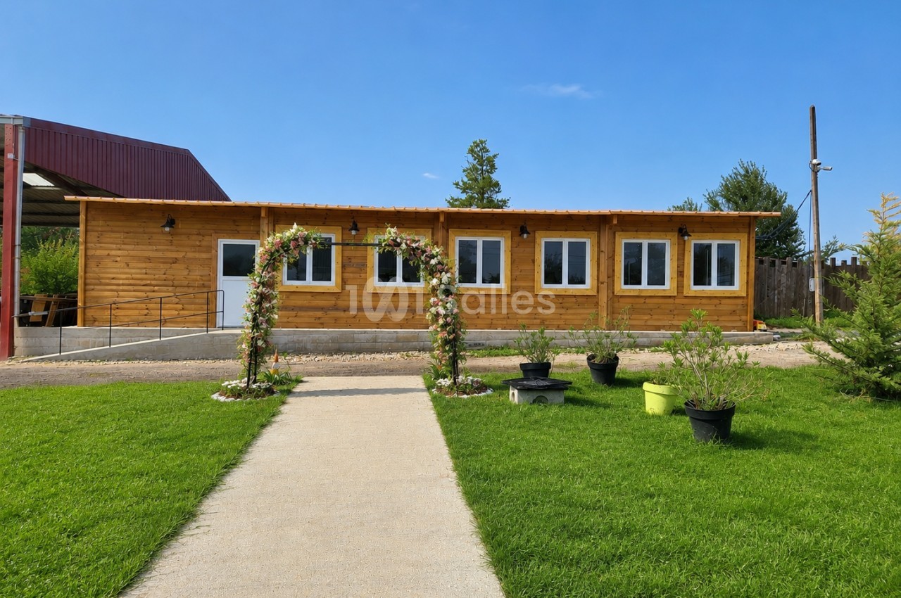 Bâtiment en bois avec six fenêtres, entrée fleurie, allée dallée et pelouse devant une structure industrielle.