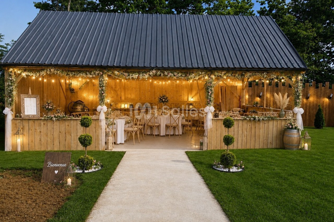 Grange en bois avec toit ardoise, intérieur illuminé aux guirlandes, tables dressées pour réception champêtre en soirée.