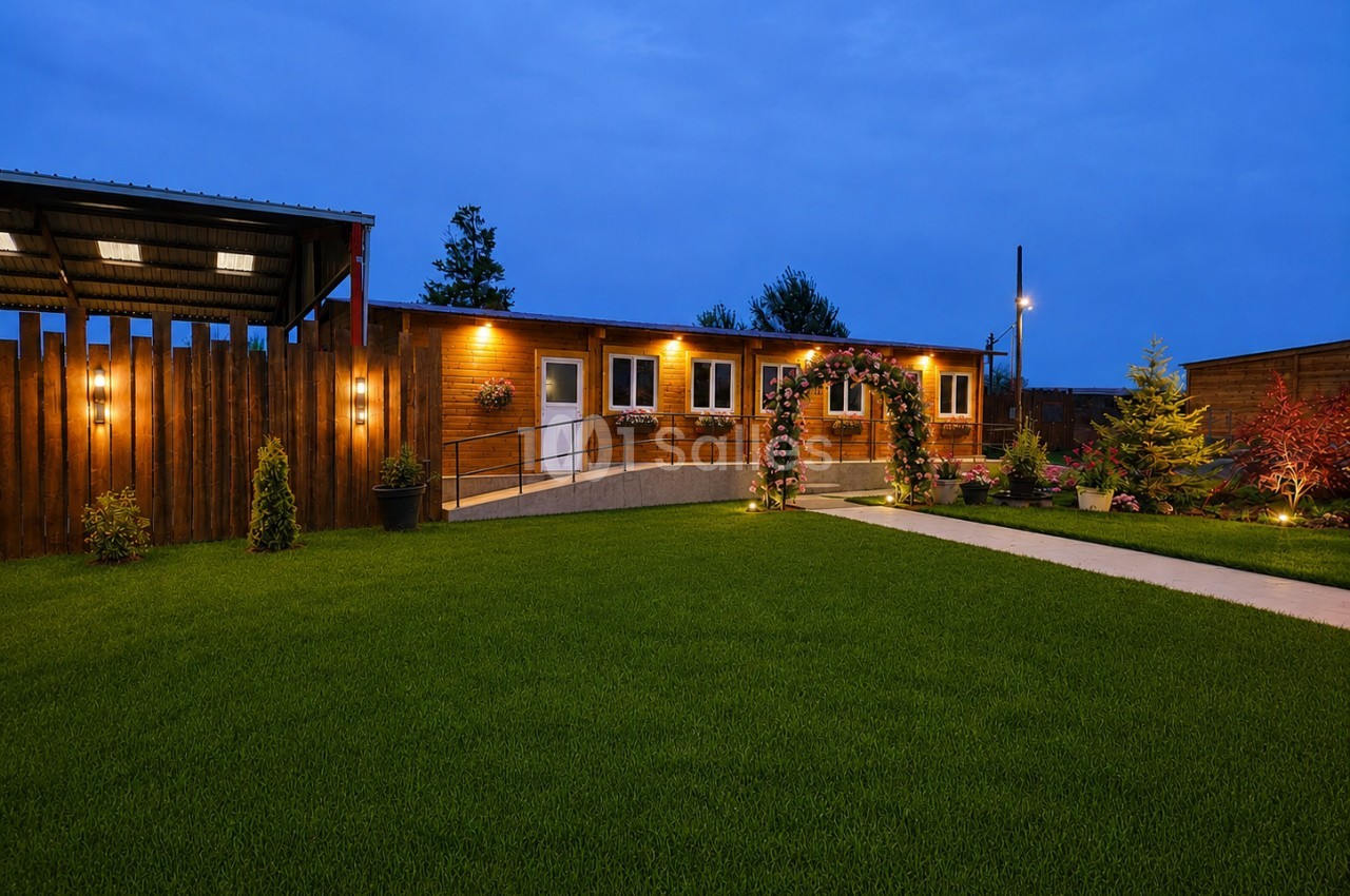 Bâtiment en bois avec éclairage nocturne, pelouse verdoyante et allée pavée encadrée de plantations paysagères.