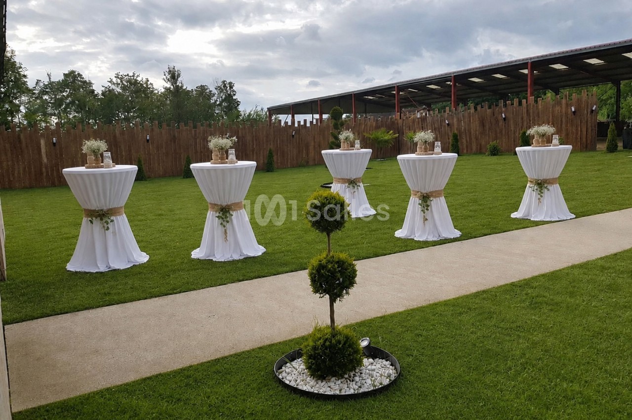 Espace extérieur gazonné avec cinq tables hautes drapées de blanc et fleurs, allée pavée, clôture bois et structure couverte en arrière-plan.